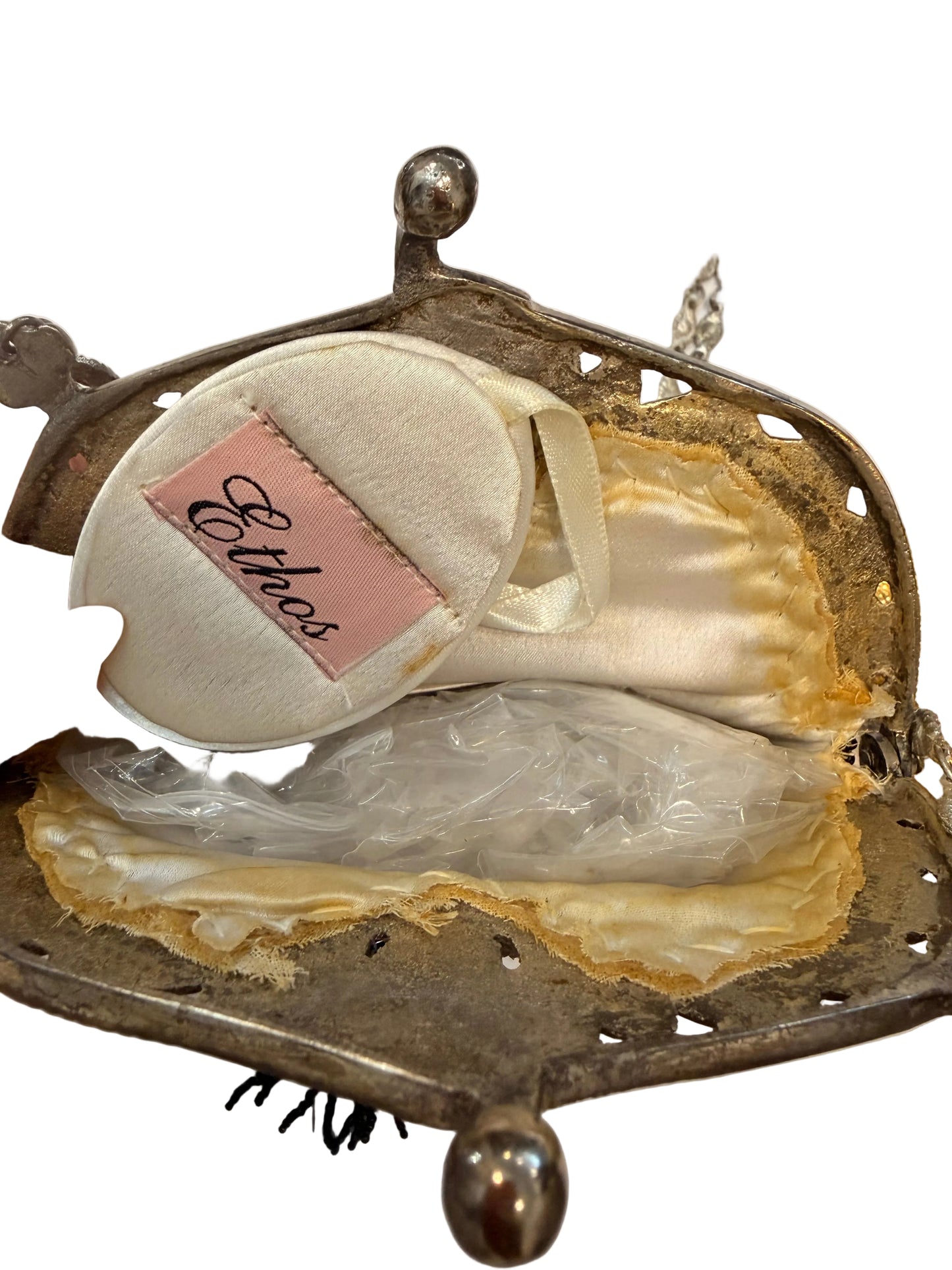 A vintage silver metal kiss-lock handbag shown open, revealing cream satin lining and a round white powder puff with a pink label that reads 'Ethos'.