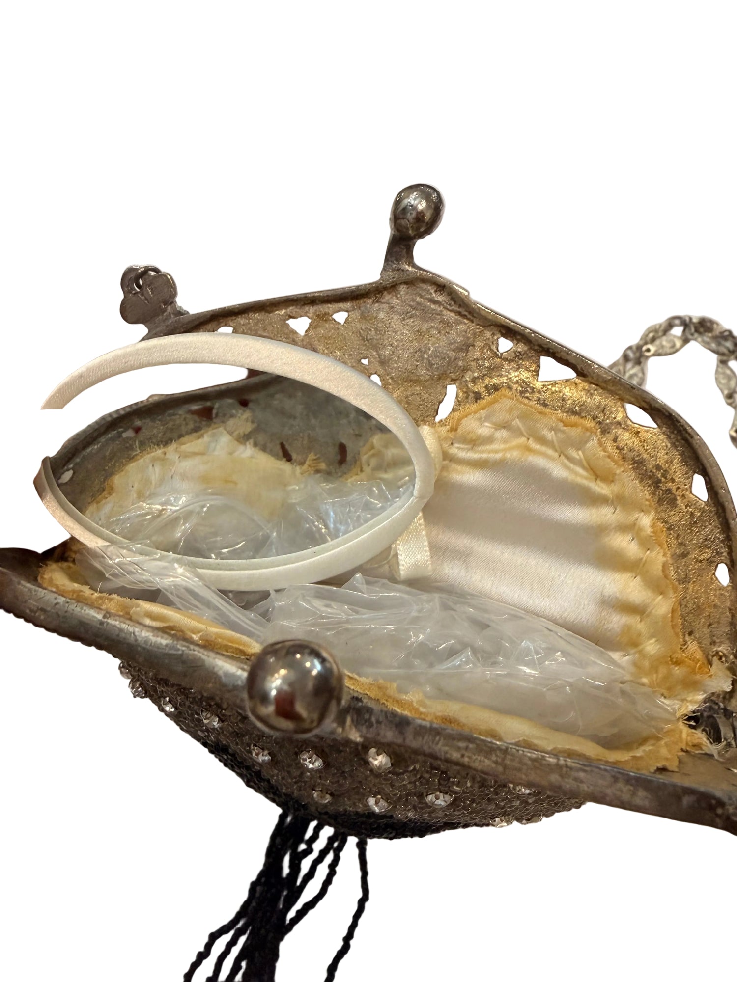 A close-up of an antique-style metallic purse or clutch with a round mirror, held open by a hand. The purse has a tarnished silver-tone metal frame with decorative ball finials, a cream satin lining, and rhinestone embellishments.
