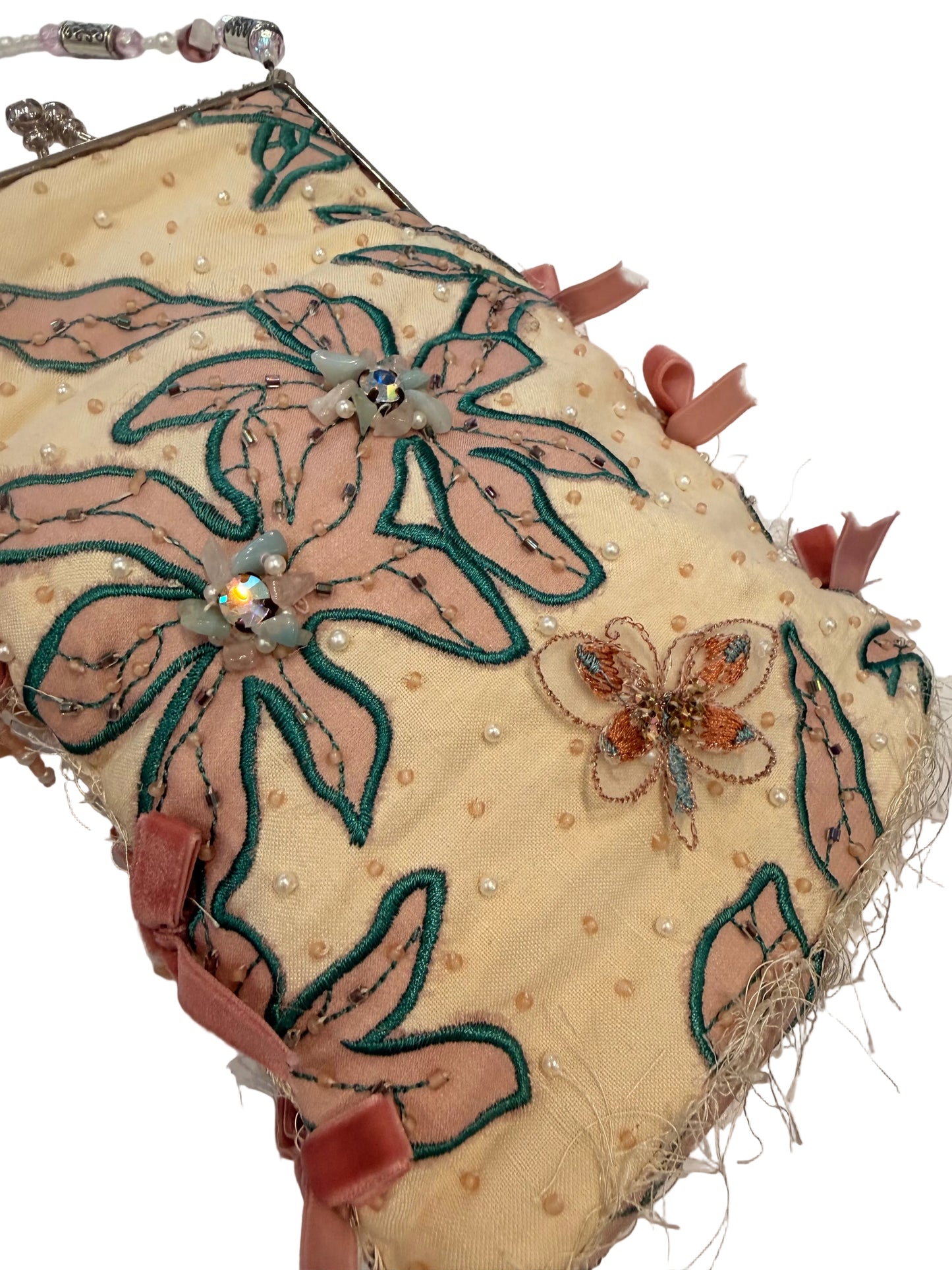 A close-up of a decorative fabric accessory, likely a small purse or pouch, featuring a cream-colored base with large embroidered floral motifs in dusty pink and dark teal-green. The piece is embellished with small pearl-like beads, a butterfly applique, and pink ribbon ties.