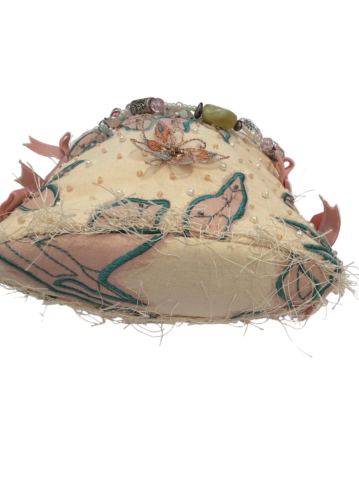 A handmade, mixed-media fabric hat with a cream base, teal embroidered lines, pink floral details, faux fur, and bead embellishments, photographed on a white surface.